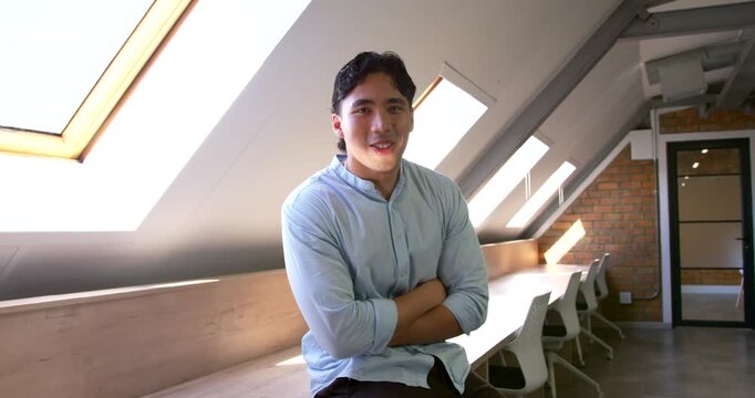 Asian man sitting on bench under skylights in loft office, reacting to off-camera cue, laughing
