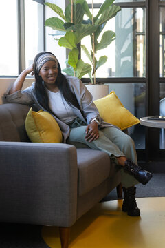 Woman sitting on gray sofa in lounge, leaning back smiling, wearing headscarf and smartwatch