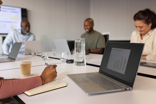 Diverse coworkers in business wear reviewing bar chart on silver laptop at table, noting notebook