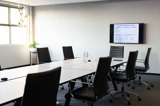 White conference table sitting in meeting room, black chairs circling, monitor showing charts