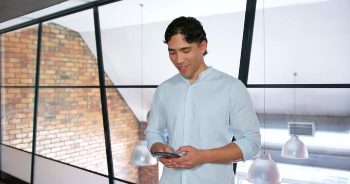 Adult man wearing light blue shirt at glass wall in office checking smartphone smiling typing reply