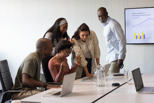 Diverse coworkers celebrating success around meeting table with woman in pink top, laptops, carafe