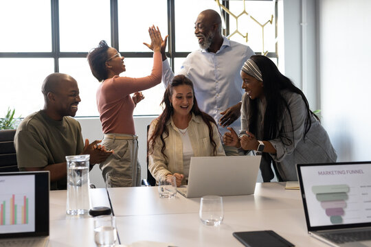 Diverse coworkers clapping and high-fiving while center woman typing on laptop at conference table