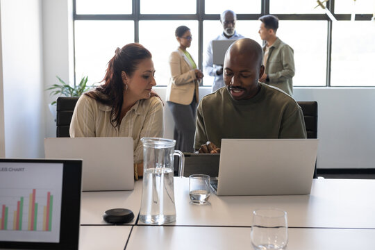 Diverse coworkers sitting at table in business casual, reviewing sales chart on monitor and laptops