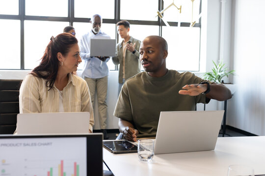Diverse coworkers discussing project in conference room with laptops, tablet, sales chart