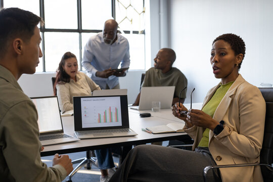 Diverse team watching woman in tan blazer speaking at meeting with laptops, sales chart, copy space