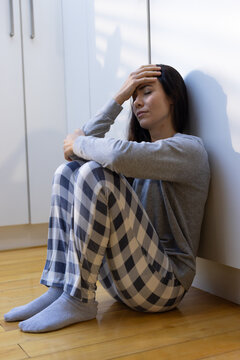 Woman sitting on hardwood floor hugging knees, leaning against white lower cabinets with sunlight