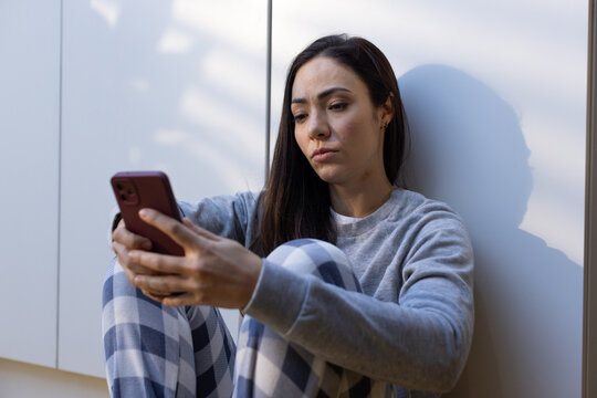 Asian woman sitting on floor by lower kitchen cabinets holding red phone, wearing grey sweatshirt