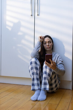 Woman sitting against white base cabinets on floor in kitchen wearing plaid pajamas, holding phone
