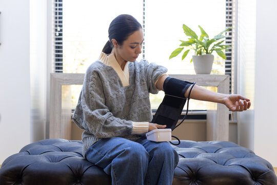 Woman sitting on ottoman by window, wearing gray sweater, adjusting blood-pressure cuff and monitor