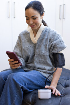 Asian woman in gray sweater sitting on ottoman at home checking phone and blood pressure cuff