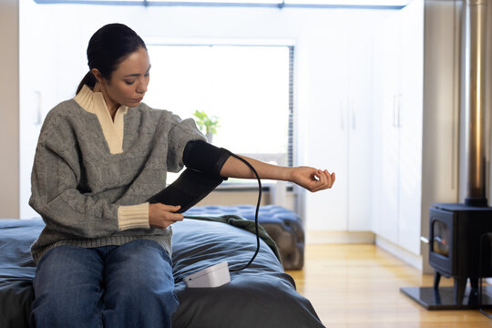 Adult Asian woman sitting on bed in bedroom, placing black blood pressure cuff with white monitor