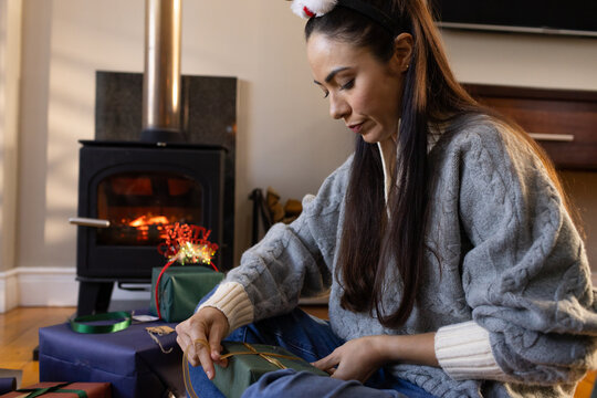 Adult female sitting hardwood floor tying ribbon on green gift box by lit stove wearing sweater