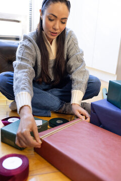 Asian woman wearing gray sweater, headband wrapping large red present on floor placing gold ribbon