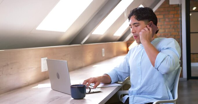 Asian man receiving call holding phone, tapping laptop and pointing at notes, managing work at desk