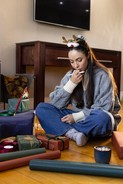 Female sitting on hardwood floor wrapping gifts in gray sweater, reindeer headband, marker, mug