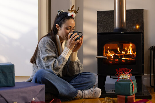 Woman sitting at home near burning stove, holding mug, wearing antler headband among wrapped gifts