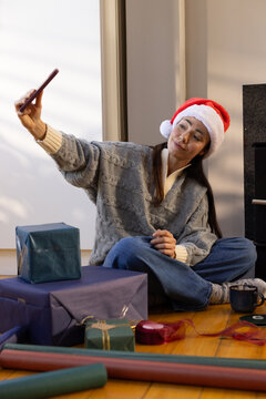 Adult woman sitting in living room wearing Santa hat taking selfie with phone among wrapped gifts
