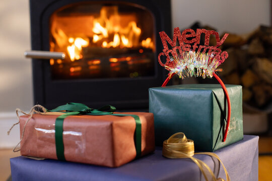 Three wrapped gifts are resting on low table near cast-iron stove, glowing flames and gold ribbon
