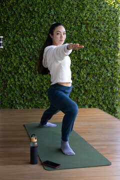 Woman practicing yoga on green mat, deck by hedge, with water bottle and phone wearing hoodie
