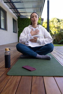 Adult woman sitting cross-legged on green mat on deck, practicing breathing with hoodie and phone