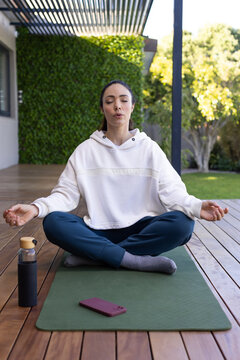 Woman sitting meditating on green mat on wooden deck, pink smartphone on mat, wearing hoodie