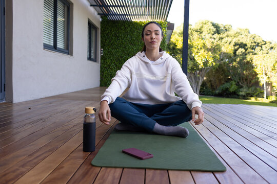 Woman sitting meditating cross-legged on yoga mat on deck, hoodie, water bottle and purple phone