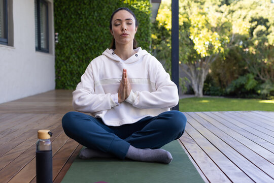 Adult woman meditating cross-legged in hoodie and sweatpants on green mat on deck with water bottle