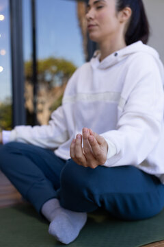 Asian woman sitting cross-legged on green yoga mat on deck, wearing white hoodie, holding mudra