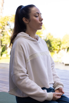 Woman sitting eyes closed on wooden deck on green yoga mat, wearing hooded pullover meditating