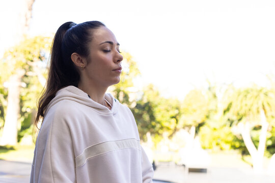 Female standing left of frame, breathing deeply in park wearing white hoodie, copy space