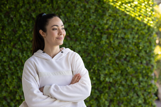 Woman standing with arms crossed wearing pullover hoodie in front of dense hedge, copy space