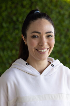 Smiling woman standing in front of green hedge, wearing white hoodie, ponytail and gold hoops