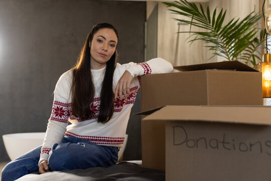 Woman wearing red patterned sweater and jeans sitting on bed sorting boxes labeled Donations