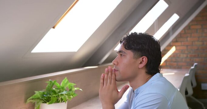 Asian man sitting at desk holding hands steepled gazing at skylight pausing by laptop, copy space