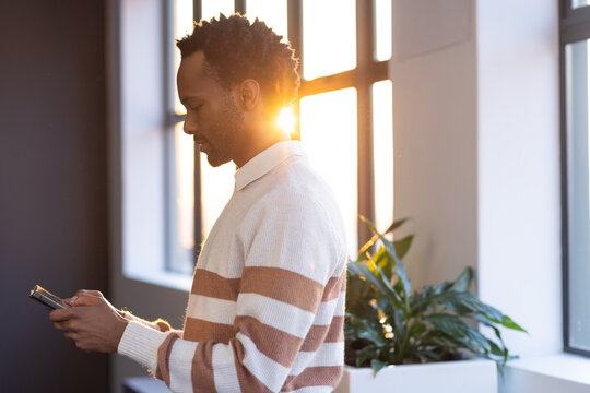 African American man in striped knit standing by grid windows in office checking phone near planter