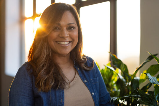 African American woman standing by paned window, sun flaring, wearing denim shirt, tan top, plants