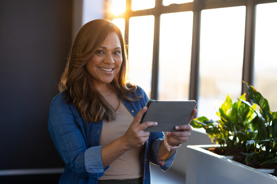 Mature African American woman standing by windows holding tablet in denim shirt and planter visible