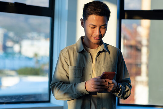 Standing mid adult Asian man wearing light overshirt and wristwatch, checking smartphone by window