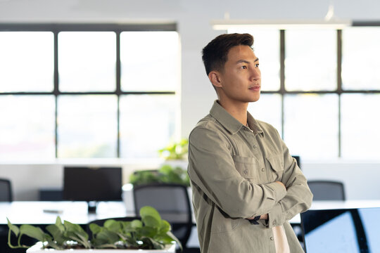 Asian man standing arms crossed wearing khaki shirt near desks and planter in office, copy space