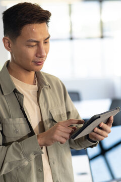 Asian man standing in classroom using tablet, khaki shirt, lanyard, chairs, windows, copy space
