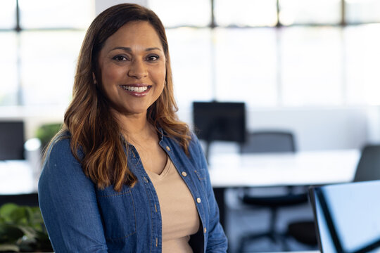 Mature African American woman smiling at camera, wearing denim shirt in modern office with monitors