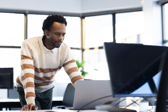 African American man leaning on desk, viewing laptop and monitor, wearing striped sweater, plant