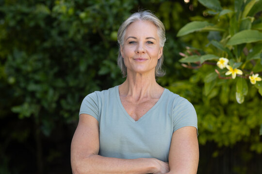 Senior female standing with arms crossed facing camera in backyard near yellow trumpet flowers