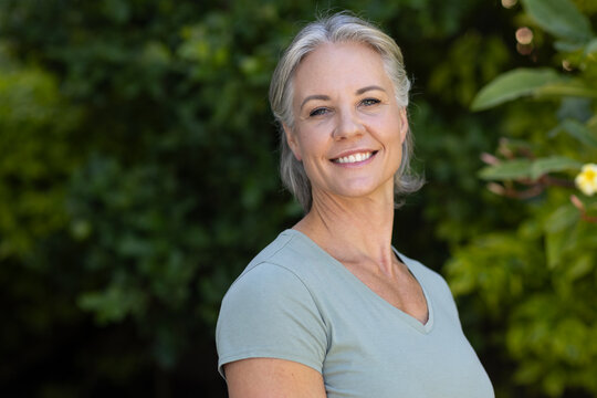 Senior female smiling, standing in garden near green shrubs wearing light green v-neck t-shirt