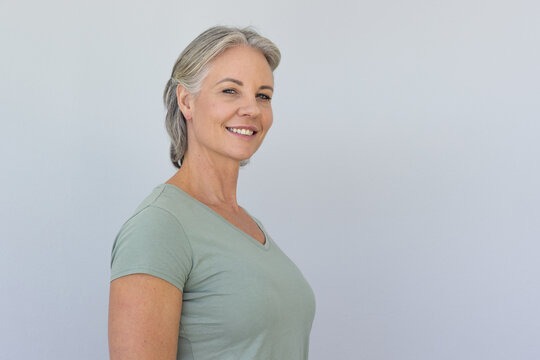 Senior woman smiling, posing in studio against pale gray wall wearing sage V-neck shirt, copy space