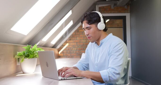 Adult man in blue shirt wearing white headset typing on laptop for work under skylights
