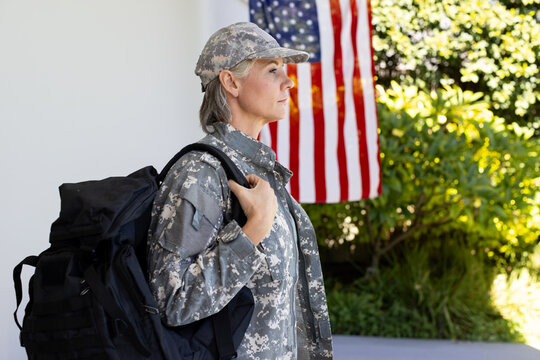 Adult woman standing in digital camo carrying black tactical pack on porch near American flag