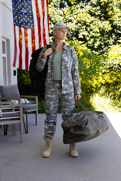 Woman in camo standing on porch holding olive duffel and black backpack near American flag