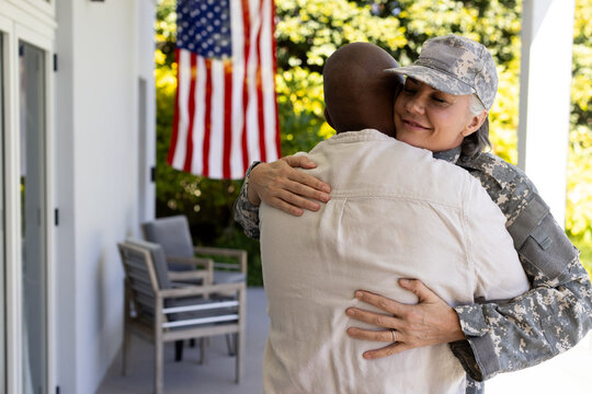 Diverse couple embracing on front porch, woman wearing camo uniform and cap, American flag nearby